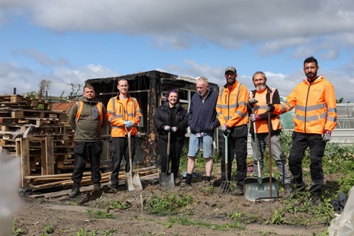 An older gentleman stands in the centre of a group of representatives from Thirteen, who are holding garden tools