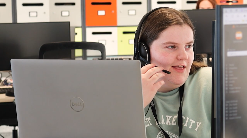 Thirteen colleague Helen at her desk working on a laptop and taking a call on her headset.