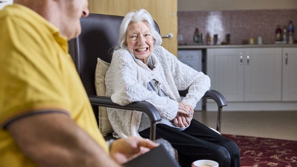 An elderly lady sat in a wheelchair smiling at a man in her home