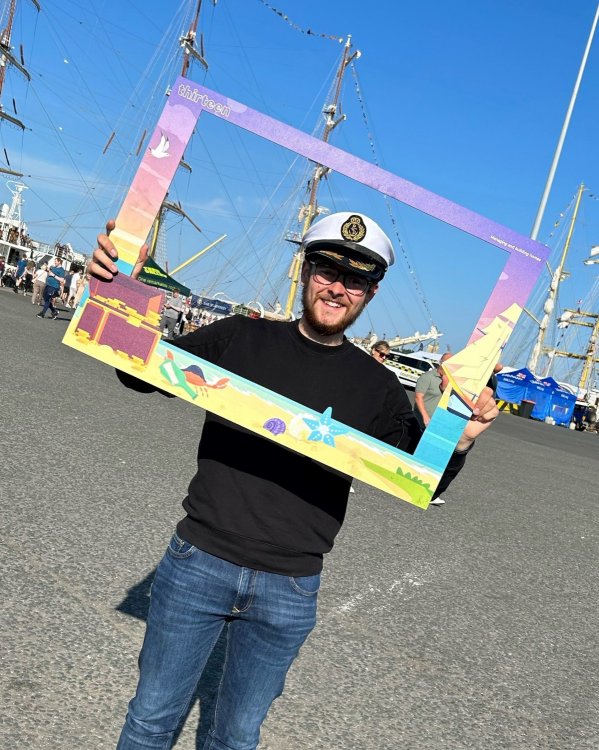 Picture of sail trainee Rhyse holding a selfie frame in front of tall ships